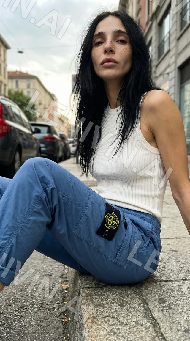 Девушка в белом топе и синих брюках Stone Island сидит на бордюре на городской улице