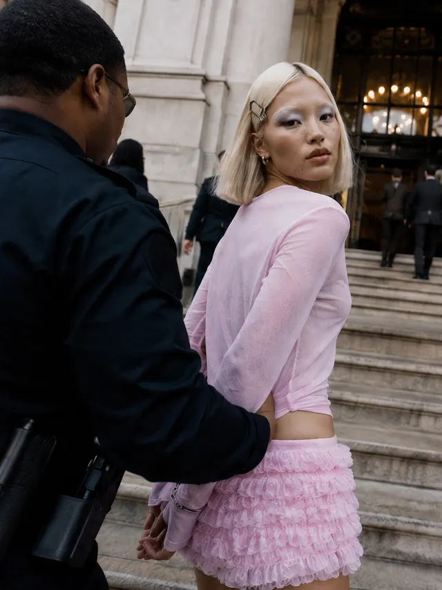 Девушка в розовом топе и рюшах стоит на ступенях, сотрудник в чёрной форме держит её за руки. Автор — Илона Врабие