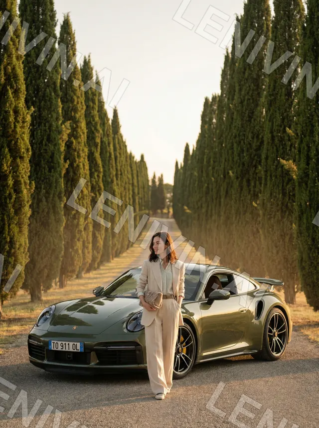 Женщина в бежевом костюме на аллее кипарисов рядом с оливковым Porsche 911 на закате