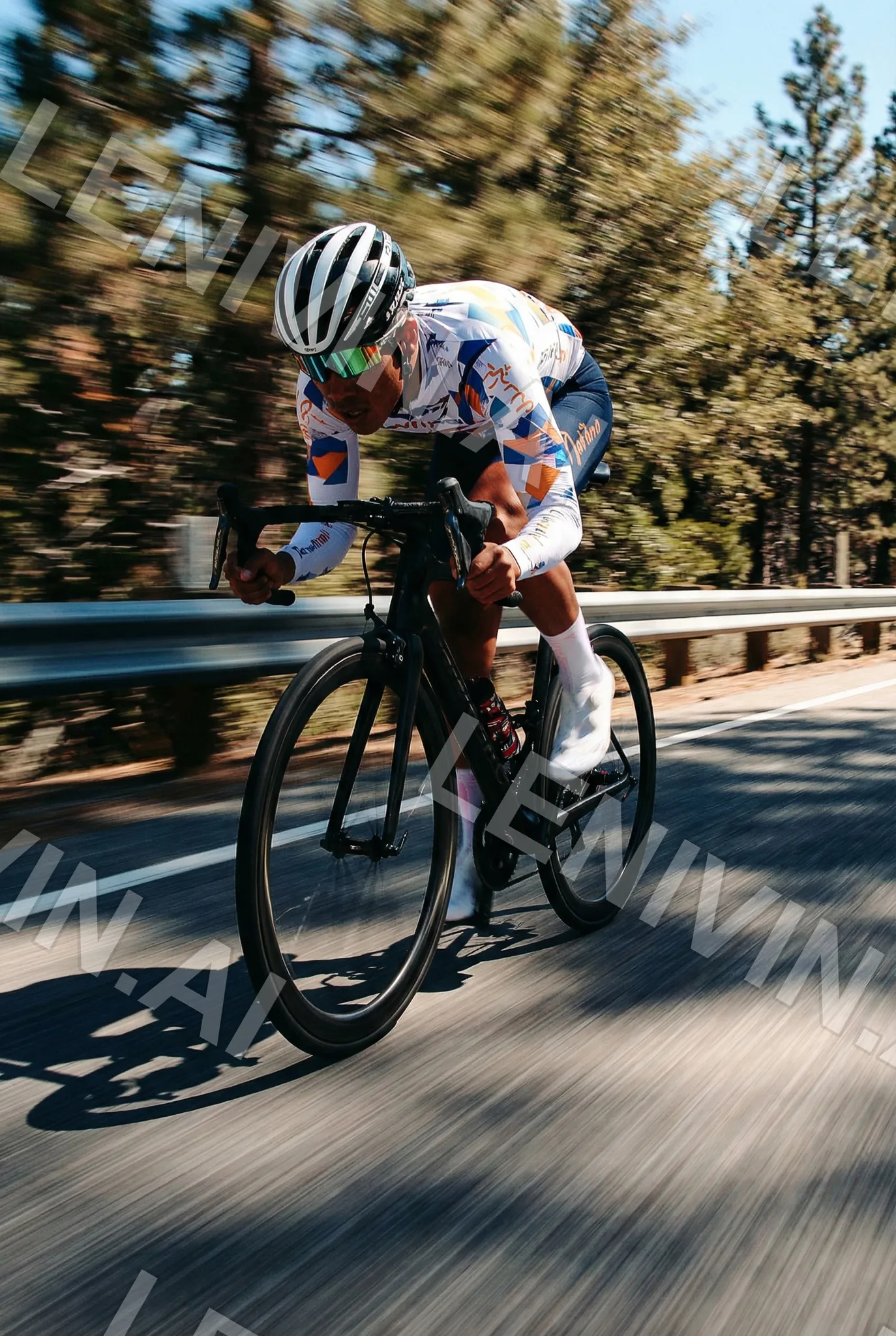 Велосипедист в яркой форме и белой каске едет по шоссе среди сосен, тень на асфальте